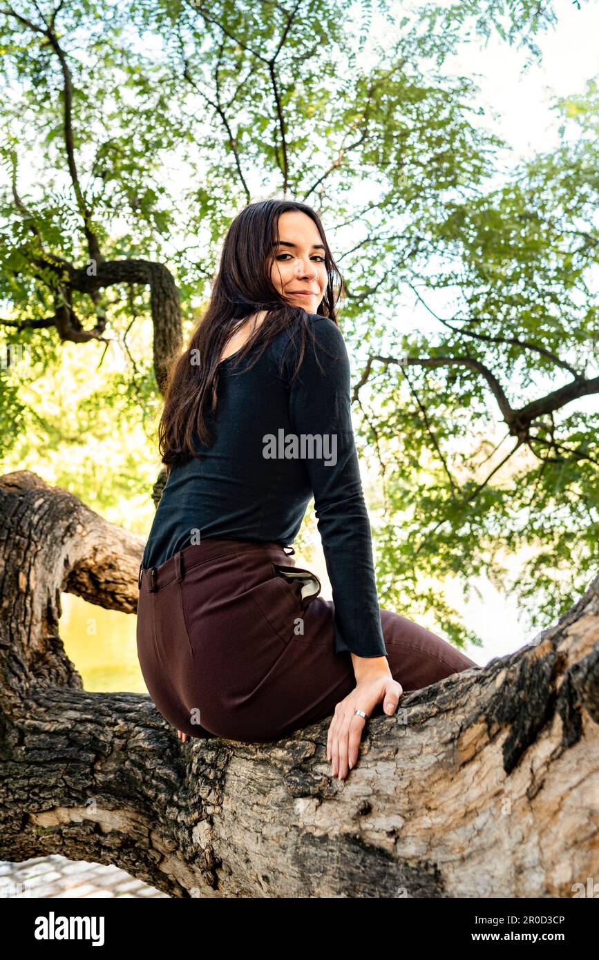 Une femme assise sur une branche d'arbre posant et regardant la caméra dans un parc public Banque D'Images