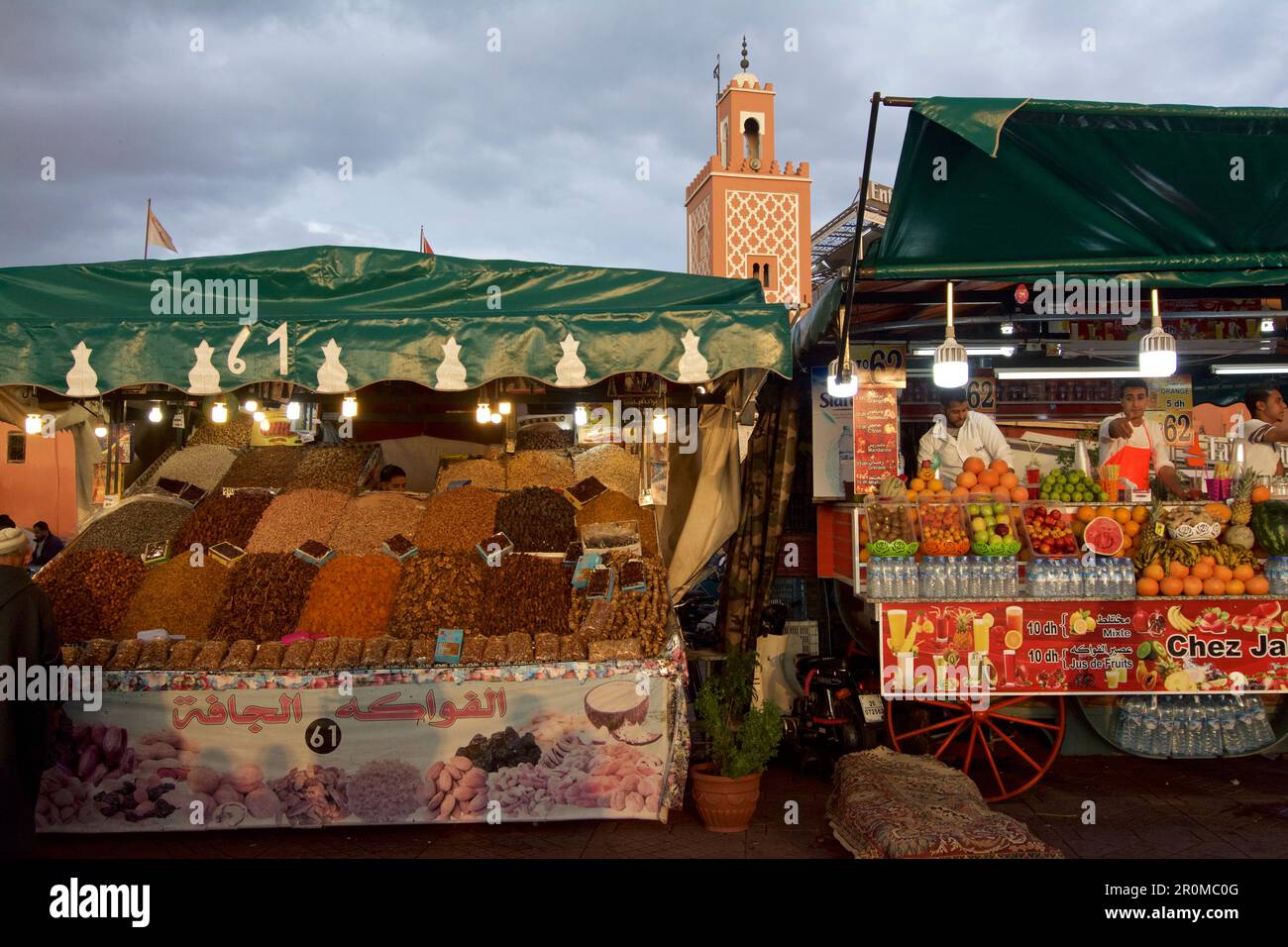 Un stand avec des fruits secs et des dattes, un stand de jus avec des fruits devant le minaret d'une petite mosquée, Jemaa El Fna, Marrakech, Maroc Banque D'Images