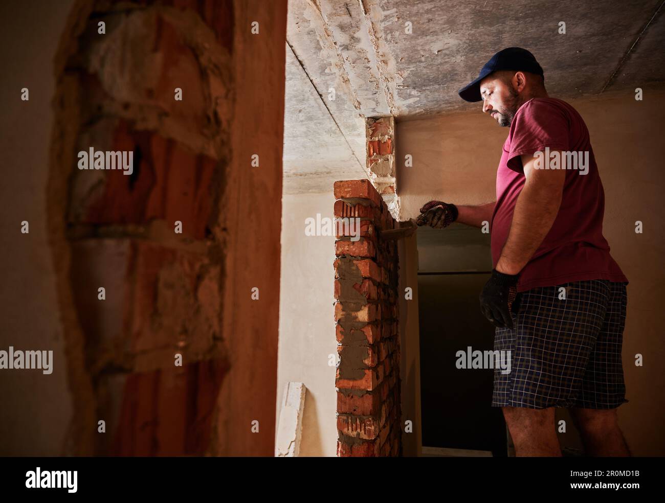 Vue latérale d'un travailleur de la maçonnerie de sexe masculin qui pose des briques dans un bâtiment en construction. Travailleur masculin appliquant du mortier de ciment sur des briques avec un outil à truelle. Concept de construction en maçonnerie. Banque D'Images
