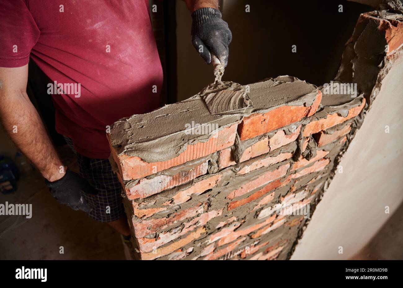 Gros plan des mains masculines dans des gants de travail portant des briques dans un bâtiment en construction. Homme bricklayer constructeur application de mortier de ciment sur les briques avec l'outil de truelle. Concept de construction en maçonnerie. Banque D'Images