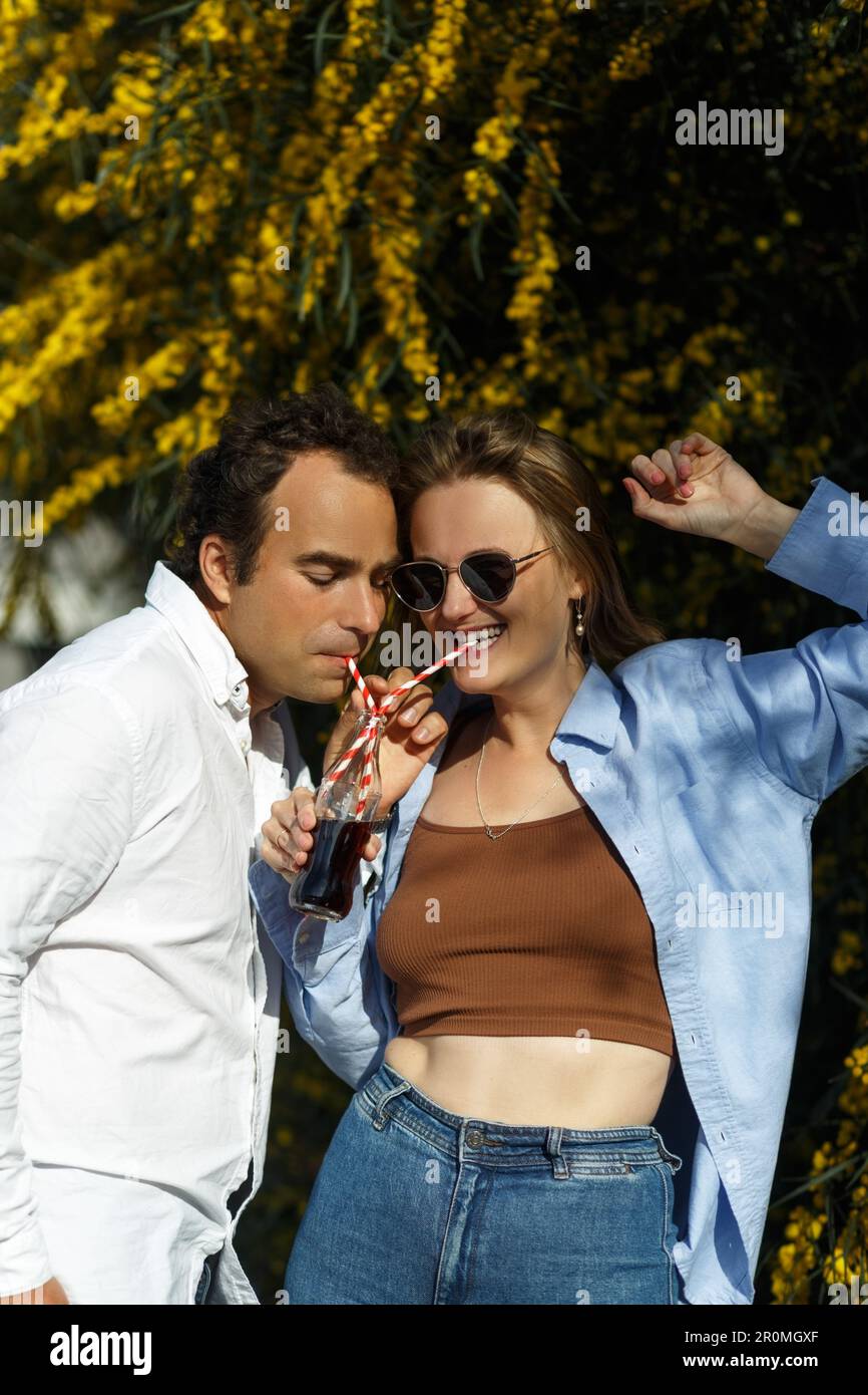 Jeune couple heureux à l'extérieur de boire une boisson froide de bouteille de verre avec de la paille. Vue latérale d'un homme et d'une femme qui boivent une boisson sans alcool et sourient. Jaune fleuri Banque D'Images