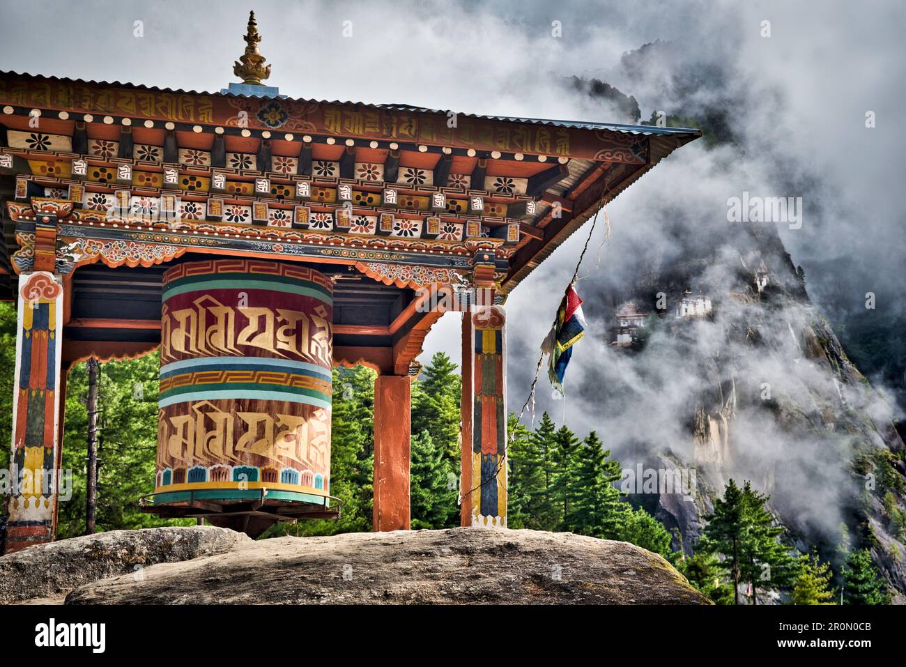 Roue de prière sur un rocher dans la forêt sur la montée au monastère Taktshang (en arrière-plan) ou Taktsang ou Tigernest, un monastère bouddhiste dans le Th Banque D'Images