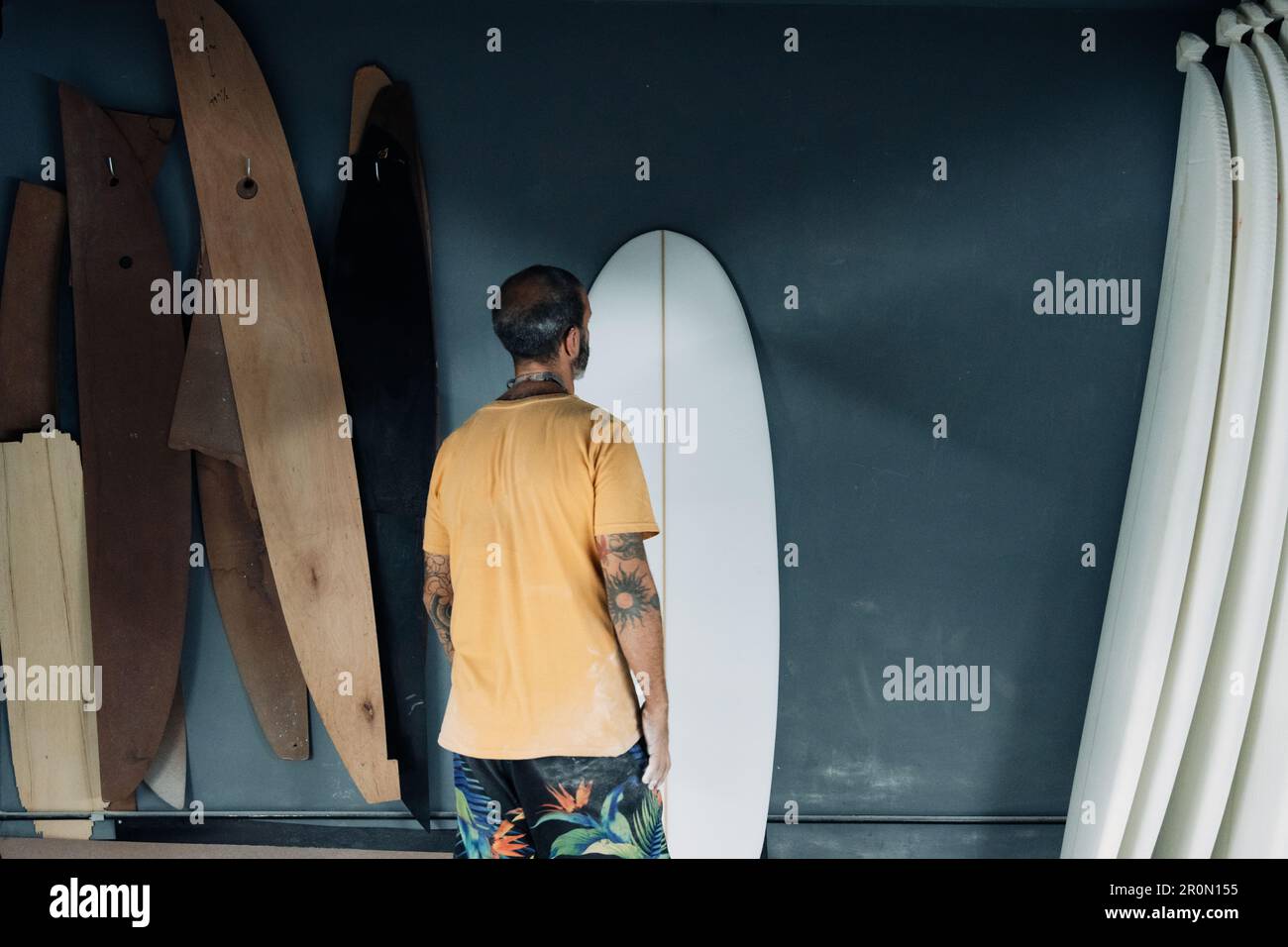 Vue arrière d'un homme maître anonyme tatoué dans des vêtements décontractés debout dans un atelier moderne et regardant la planche de surf tout en vérifiant la qualité du travail Banque D'Images Vue arrière d'un homme maître anonyme tatoué dans des vêtements décontractés debout dans un atelier moderne et regardant la planche de surf tout en vérifiant la qualité du travail Banque D'Images
