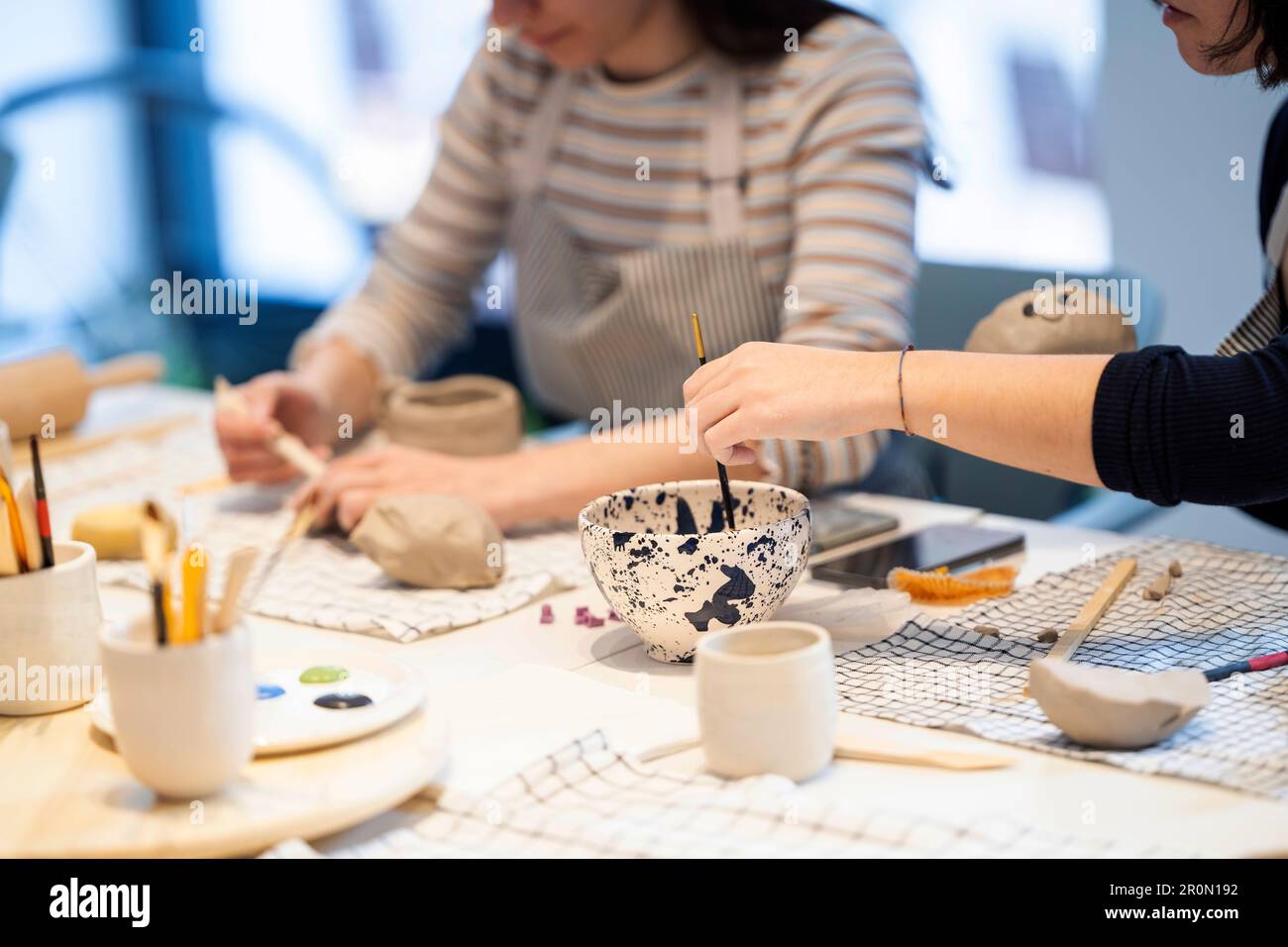 Des artistes féminins anonymes en tabliers peignant sur des tasses en céramique tout en travaillant ensemble dans un studio de poterie créatif Banque D'Images