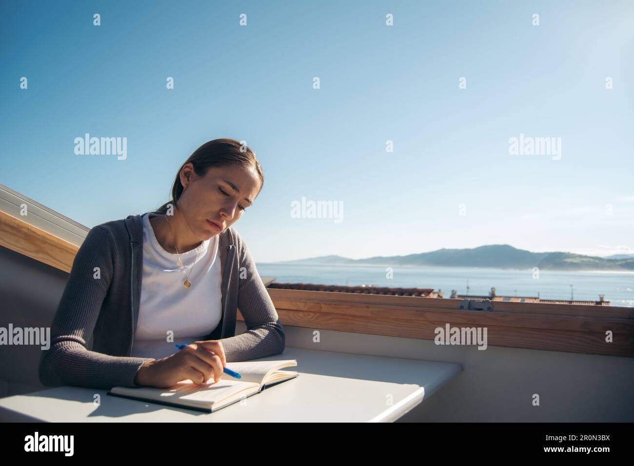 Jeune étudiante réfléchie dans des vêtements décontractés assis à table et écrivant des informations dans le bloc-notes à des paysages pittoresques et pensant à des idées Banque D'Images