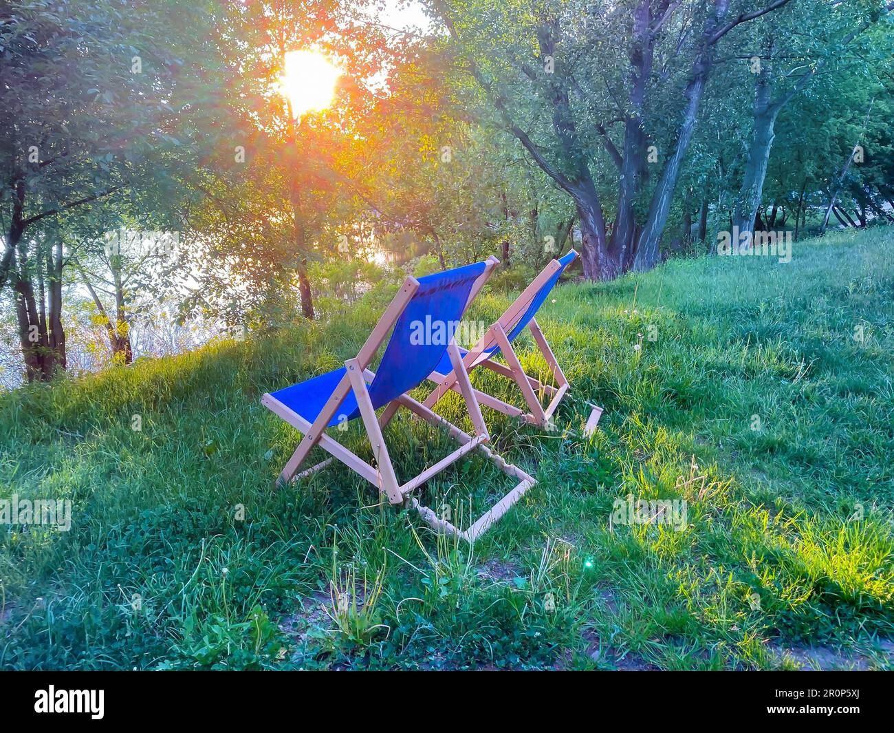 Chaises longues au coucher du soleil. deux chaises longues bleues au bord du lac entourées d'herbe et d'arbres au coucher du soleil. Détendez-vous avec un narguilé le soir en ville Banque D'Images