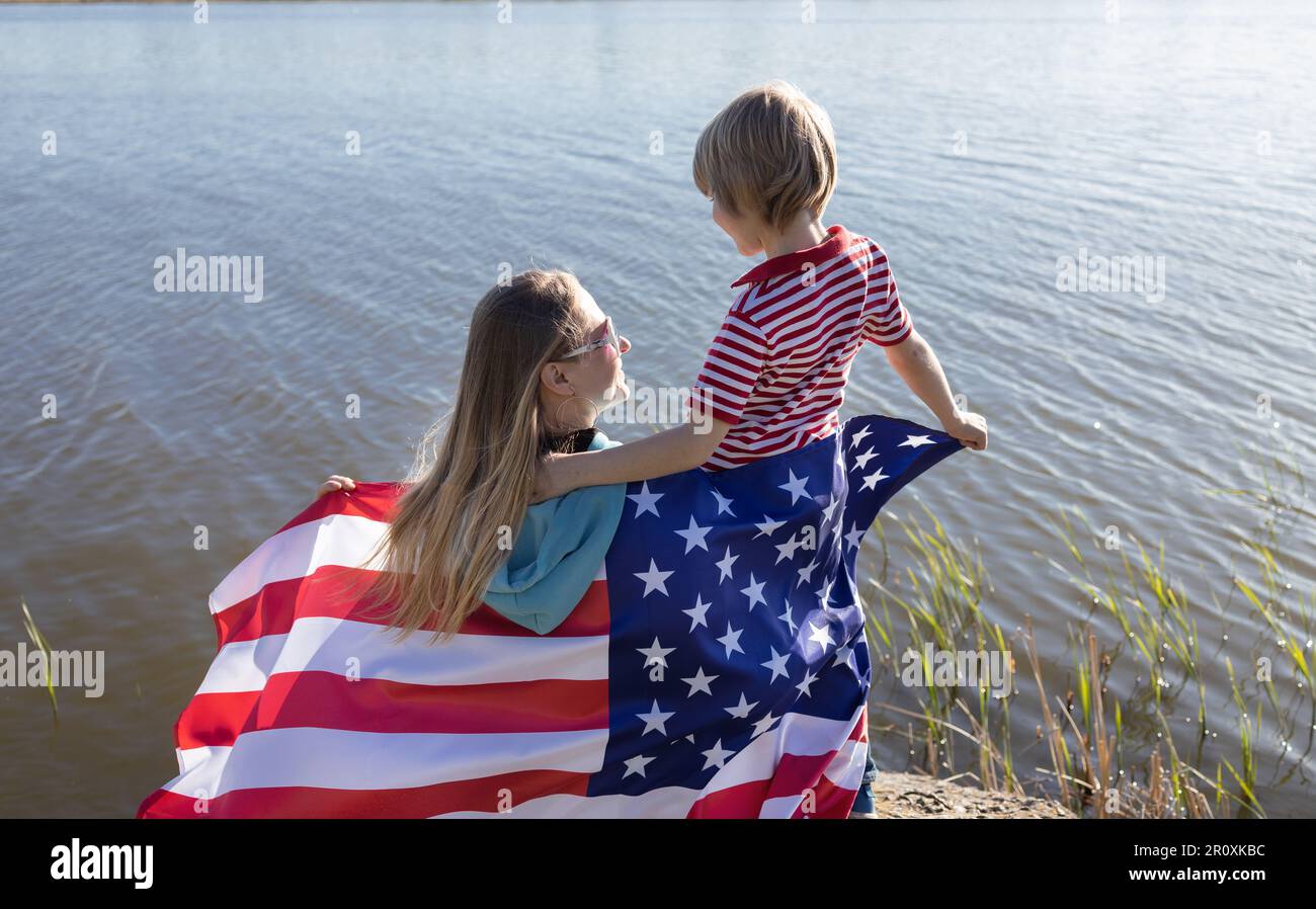 mère et enfant heureux tenant le drapeau américain derrière leur dos, assis sur le bord du lac. Liberté, identité nationale. Journée de l'indépendance des États-Unis Banque D'Images