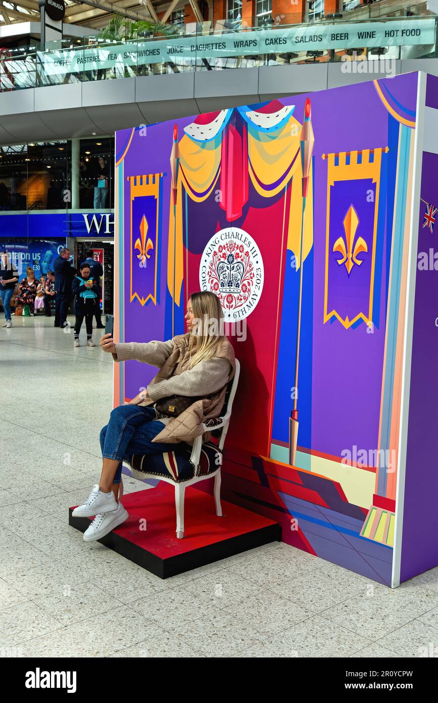 Une jeune femme blonde attirante prenant un selfie tout en étant assise devant un décor célébrant le couronnement du Roi Charles Waterloo Station UK Banque D'Images