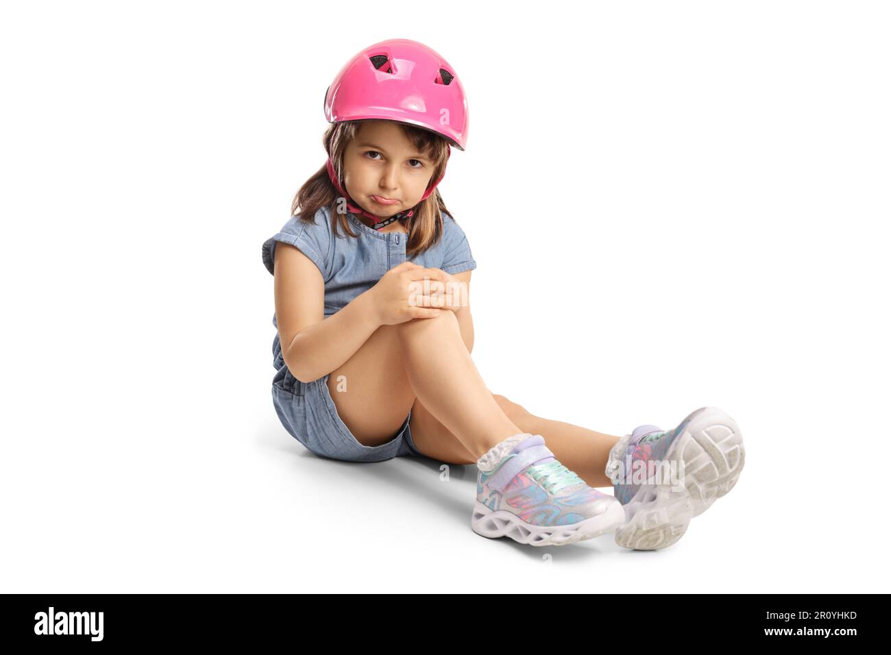 Triste petite fille avec un casque assis sur le sol et tenant son genou isolé sur fond blanc Banque D'Images