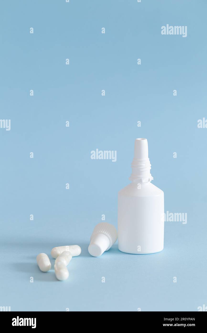 Un flacon de spray nasal avec le bouchon retiré et quelques capsules de médicament placées à côté. Produits blancs sur fond bleu. Verticale. Banque D'Images