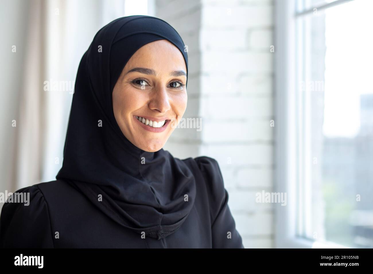Gros plan. Portrait d'une jeune belle musulmane dans un hijab noir regardant avec assurance et souriant à l'appareil photo, debout à l'intérieur près de la fenêtre. Banque D'Images