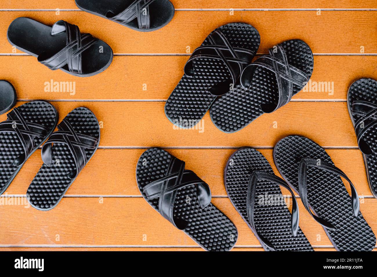 Vue de dessus de plusieurs paires de sandales noires classiques sur fond de bois peint orange sur le patio. De nombreuses paires de tongs en caoutchouc à côté de la piscine ouverte Banque D'Images