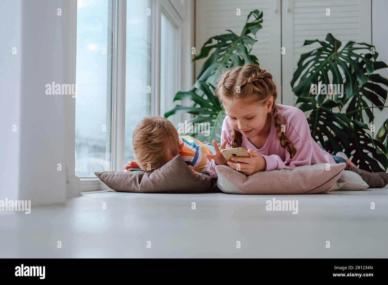 Les enfants sont au sol de la fenêtre et regardent des dessins animés sur leur téléphone. Le frère et la sœur jouent à des jeux sur le téléphone. Banque D'Images