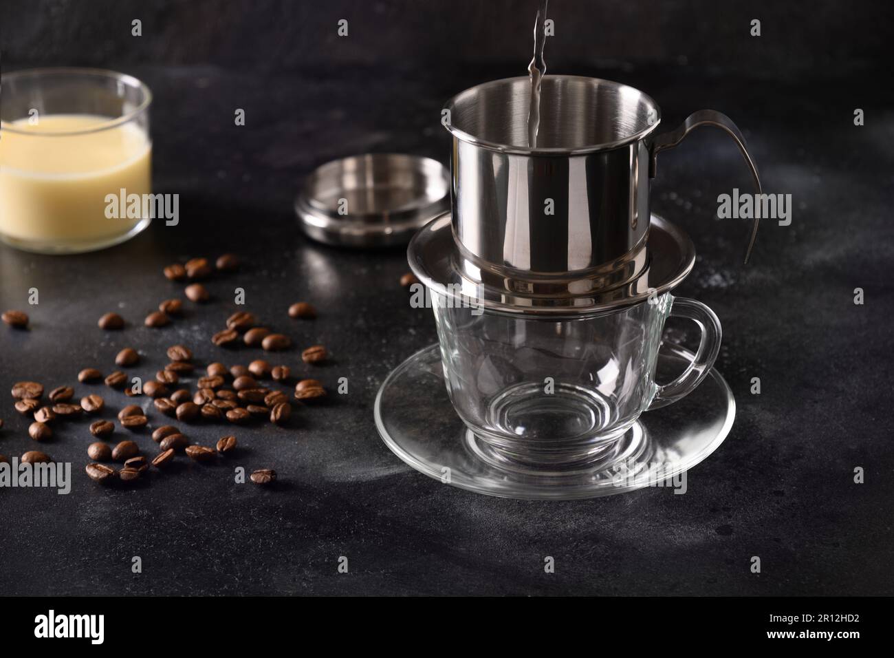 Verser de l'eau chaude dans le filtre goutte-à-goutte pour préparer du café vietnamien avec du lait concentré sucré dans une tasse en verre sur fond noir. Gros plan. Banque D'Images