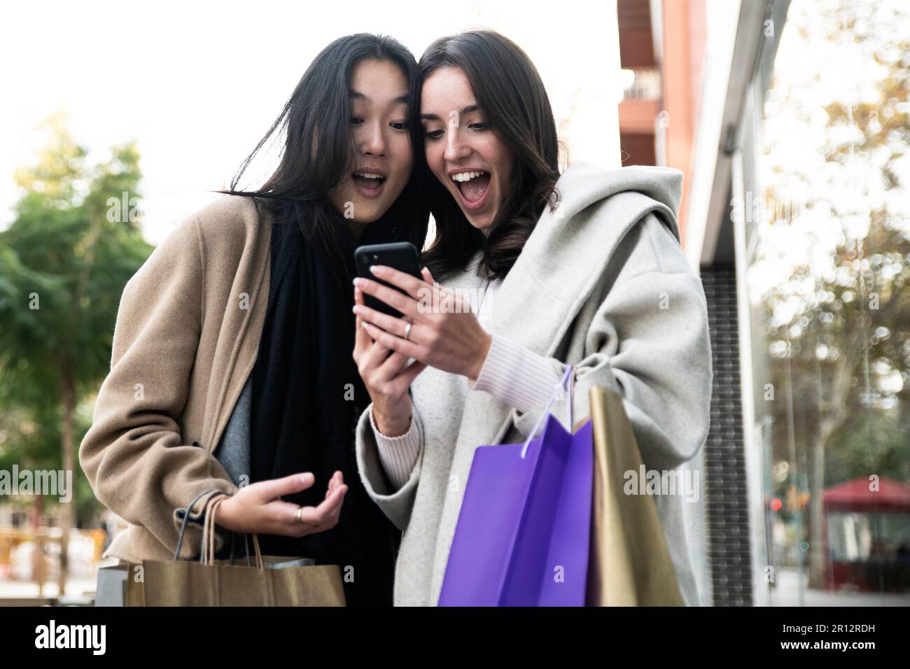 Deux jeunes femmes surprennent, belles, portant des sacs à provisions et un smartphone. Des amis joyeux émerveillés par son téléphone portable. Banque D'Images