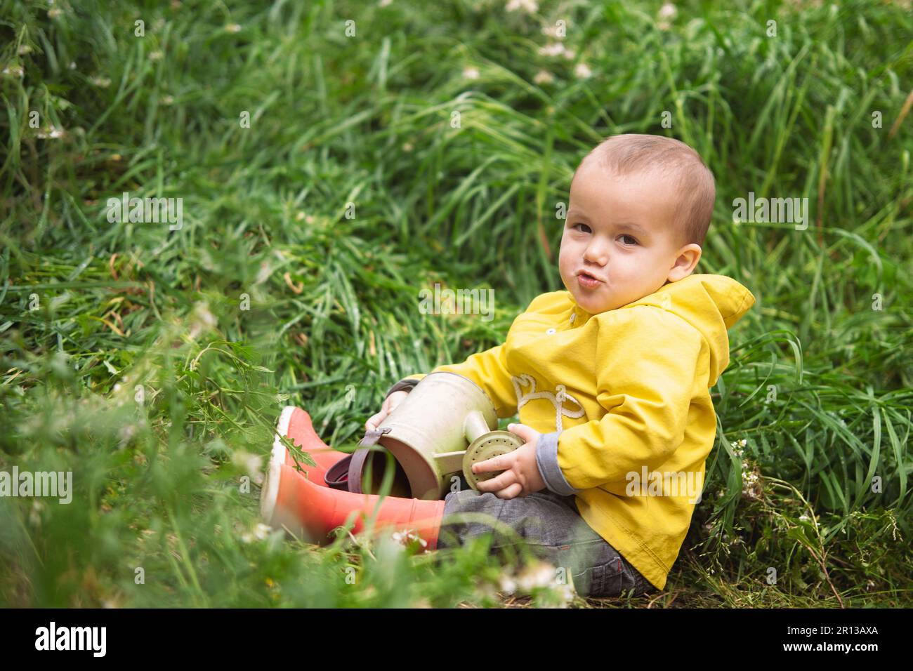 Un garçon d'un an avec une veste jaune et des bottes de pluie rouges est assis au milieu d'un pré vert, tenant un arrosoir entre ses mains. Banque D'Images