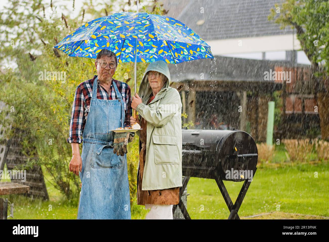 Un homme et une femme âgés font des visages déçus en se trouvant sous un parapluie devant leur barbecue encore fumeur pendant un événement de campagne, ca Banque D'Images