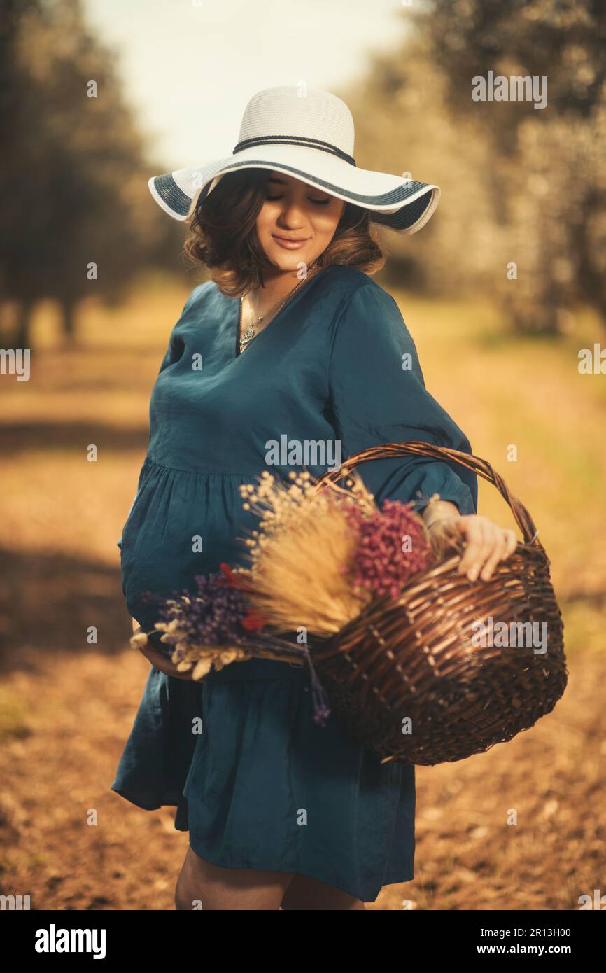 Une femme enceinte avec un chapeau blanc robe bleue et des fleurs sèches mousquetées dans un champ d'olivier à faible profondeur de champ. Banque D'Images