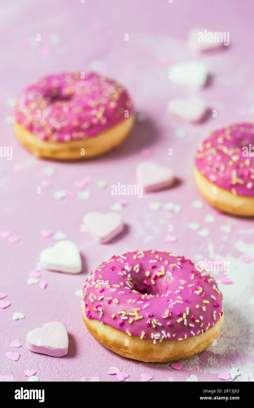 Beignets, Berliner, Krapfen avec glaçage au sucre rose sur fond texturé rose, décoré de sucre rose et blanc et de coeurs en papier, vertical Banque D'Images