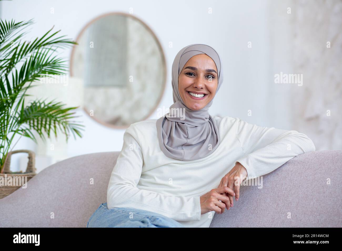 Portrait d'une jeune femme d'affaires d'origine orientale dans le hijab assis sur un canapé à la maison et regardant l'appareil photo avec assurance et sourire. Banque D'Images