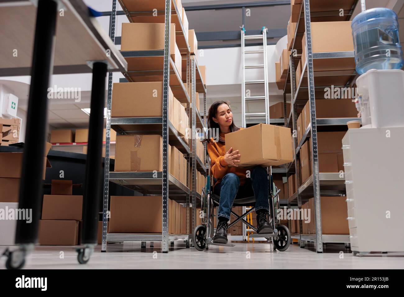 Le responsable d'entrepôt en fauteuil roulant vérifie la qualité de l'emballage des colis dans la salle de stockage. Opérateur logistique transportant une boîte en carton tout en gérant les stocks dans un magasin pour personnes handicapées Banque D'Images