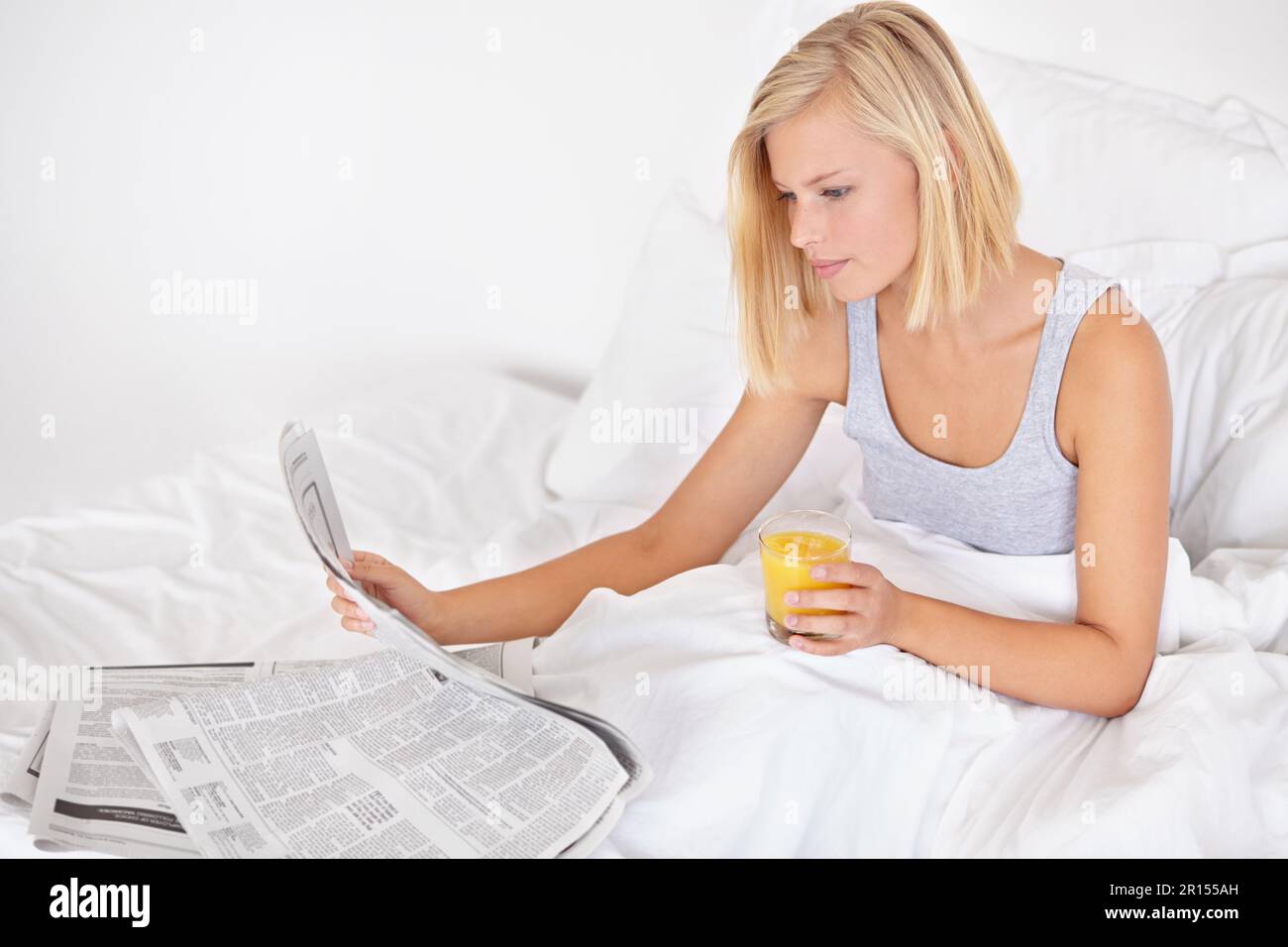 Rattraper son retard. Une jolie jeune femme lisant le journal au lit et appréciant un verre de JO. Banque D'Images