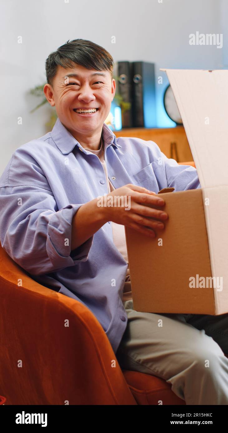 Un homme asiatique heureux déballant le colis de livraison à la maison.  Sourire satisfait chinois gars shopper en ligne client ouverture carton  réception achat cadeau par expédition rapide par la poste. Tir
