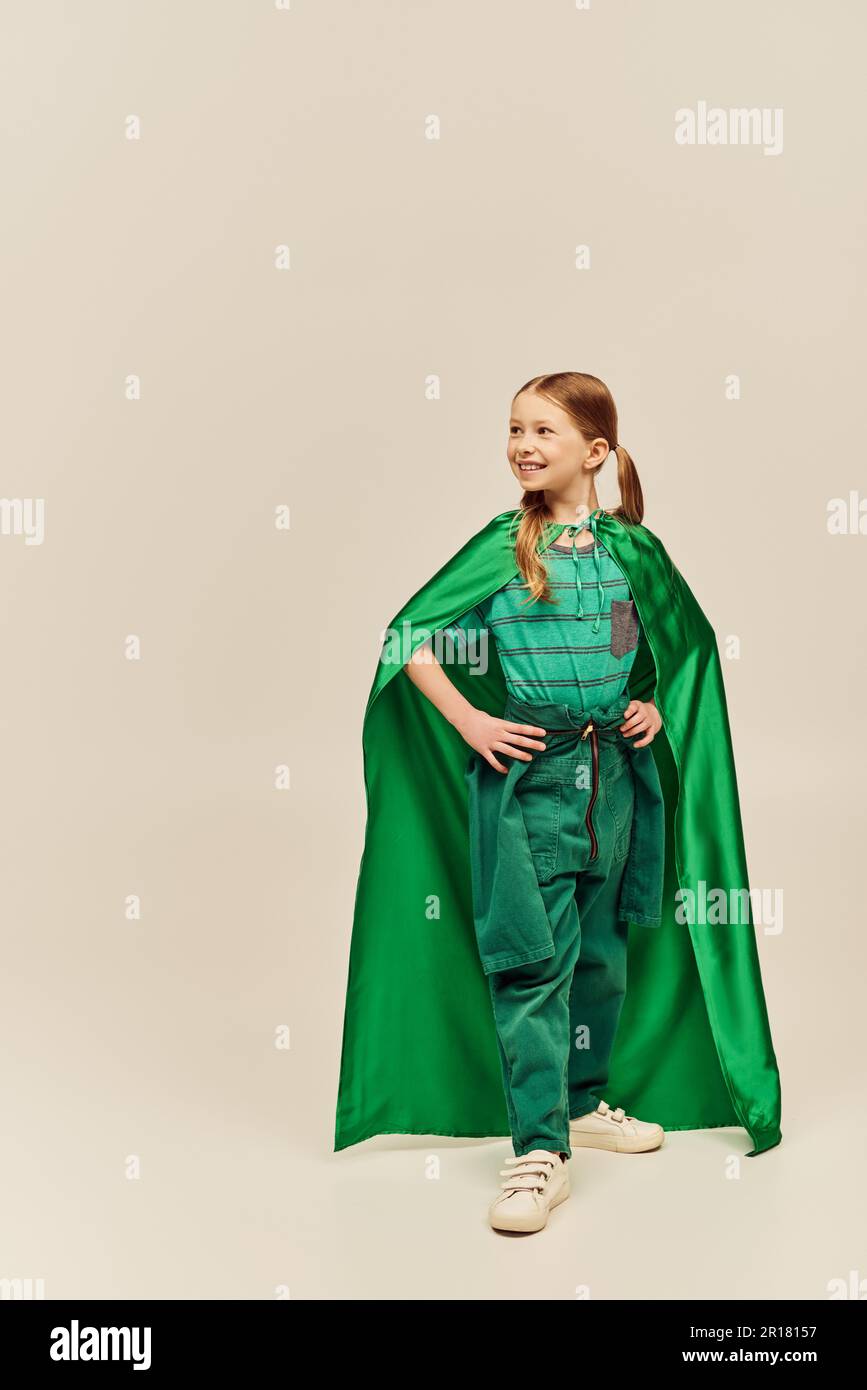 Fille souriante en costume de super-héros vert avec un manteau portant un  pantalon et un t-shirt et debout avec les mains sur les hanches tout en  célébrant les enfants internationaux Photo Stock -