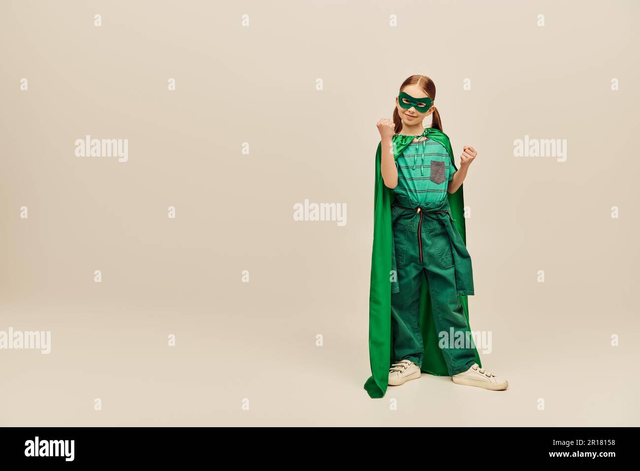 Puissante fille en costume vert super-héros avec le manteau et le masque sur le visage, portant le pantalon et le t-shirt et debout avec les poings serrés tout en célébrant le Chi Banque D'Images