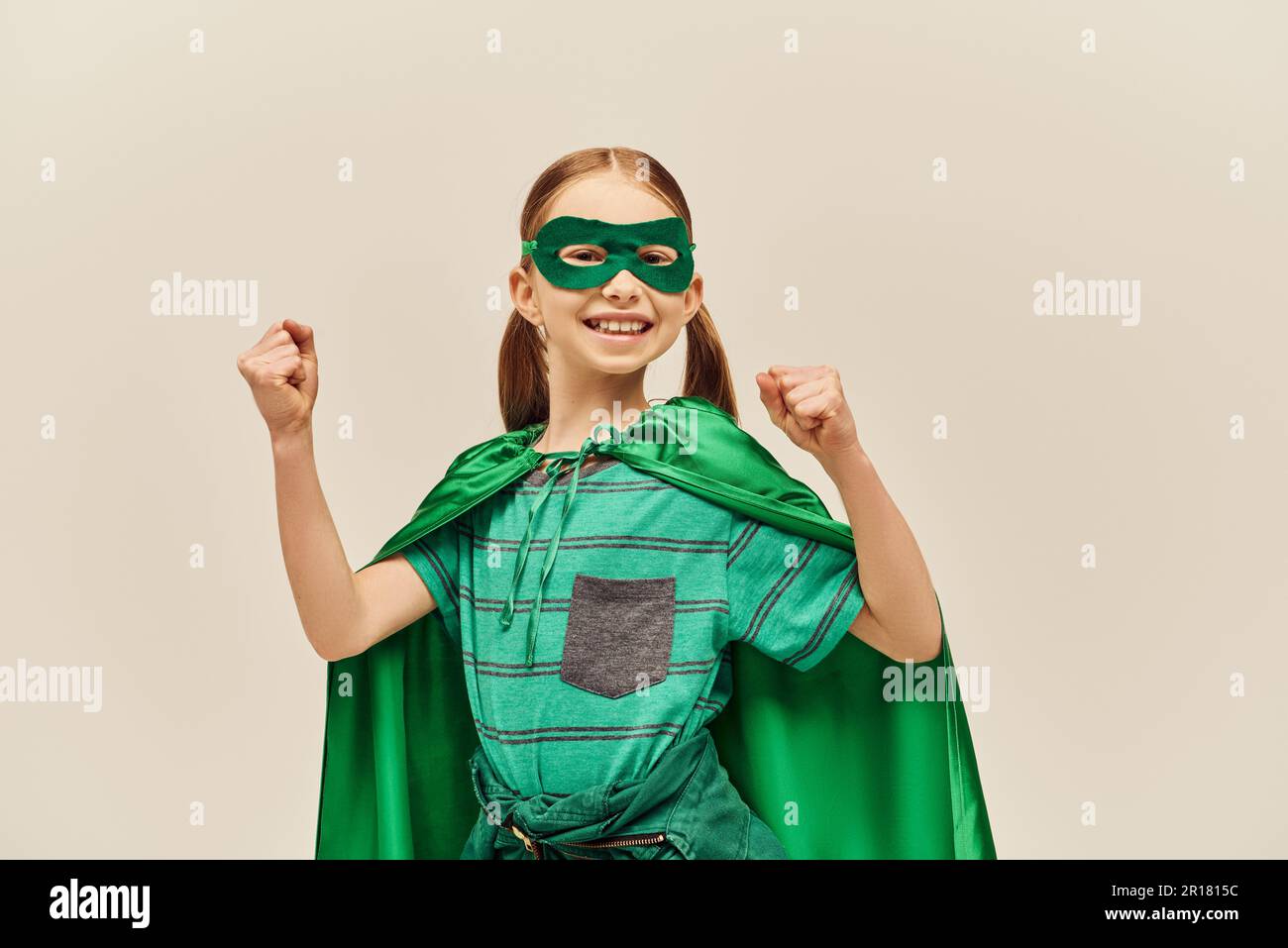 Puissante fille en costume de super-héros vert avec le manteau et le masque sur le visage, souriant et debout avec les poings serrés tout en célébrant la protection de l'enfant du monde Banque D'Images