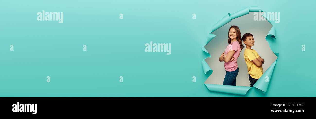 Des amis préadolescents multiethniques heureux croisant les bras et regardant la caméra tout en célébrant la journée mondiale de protection des enfants près du trou dans le papier bleu backgrou Banque D'Images