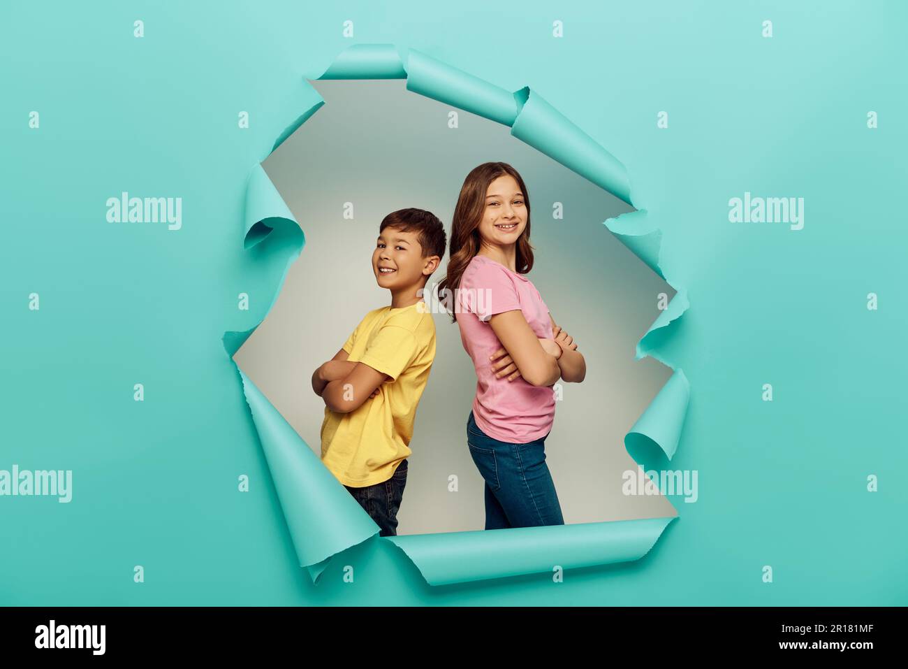 Souriant, préadolescents interraciaux dans des t-shirts colorés croisant les bras tout en se tenant dos à dos et célébrant la journée de protection de l'enfant derrière le trou dans bl Banque D'Images