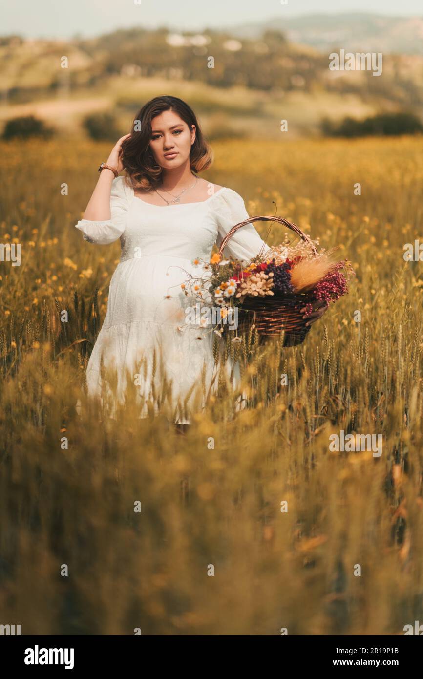 Une femme enceinte vêtue d'une robe blanche porte un panier de fleurs  sèches dans un champ de blé doré, incarnant l'esprit de la Fête des mères  Photo Stock - Alamy