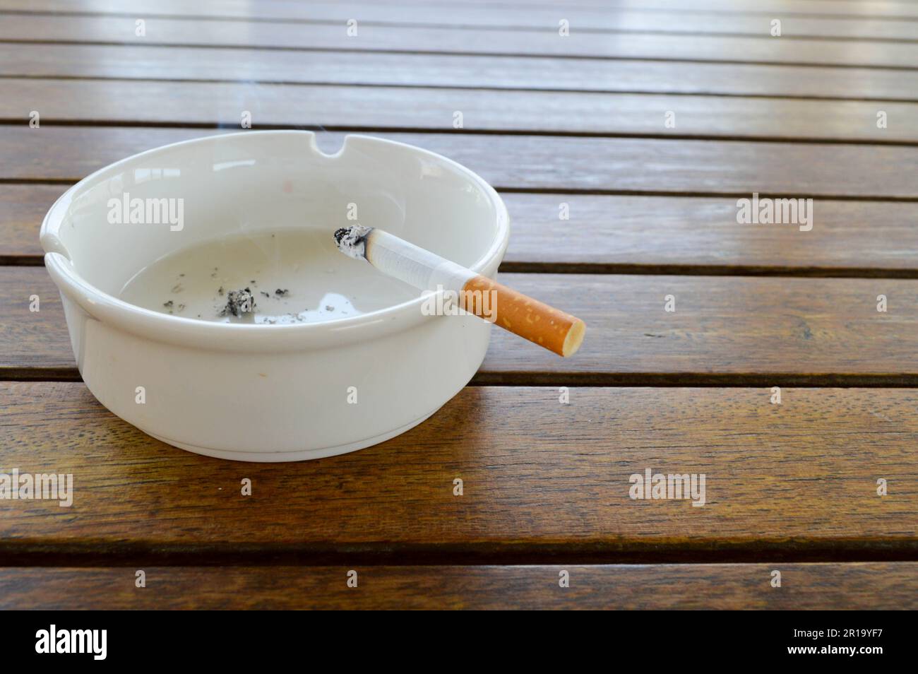 Un cendrier blanc en céramique avec une cigarette et une cendre avec un briquet noir pour fumer est posé sur une table en bois dans un café. Banque D'Images