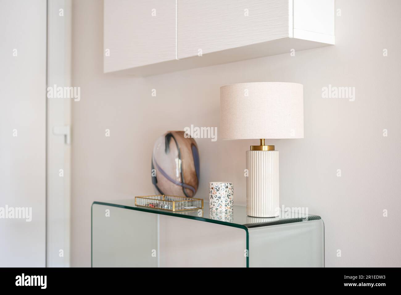 Vue rapprochée d'une console en verre avec accessoires et d'une lampe avec une armoire blanche suspendue dans le couloir ou le salon. Décoration intérieure moderne Banque D'Images