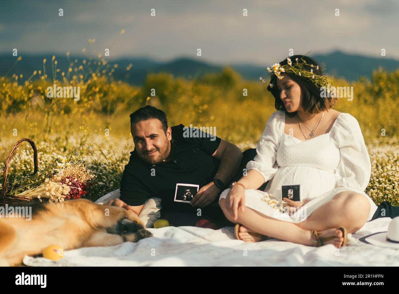 Sur une couverture de pique-nique blanche, une femme enceinte et son mari partagent un moment de tendreté avec l'image échographique de leur bébé, tandis que leur sympathique Golden Retr Banque D'Images