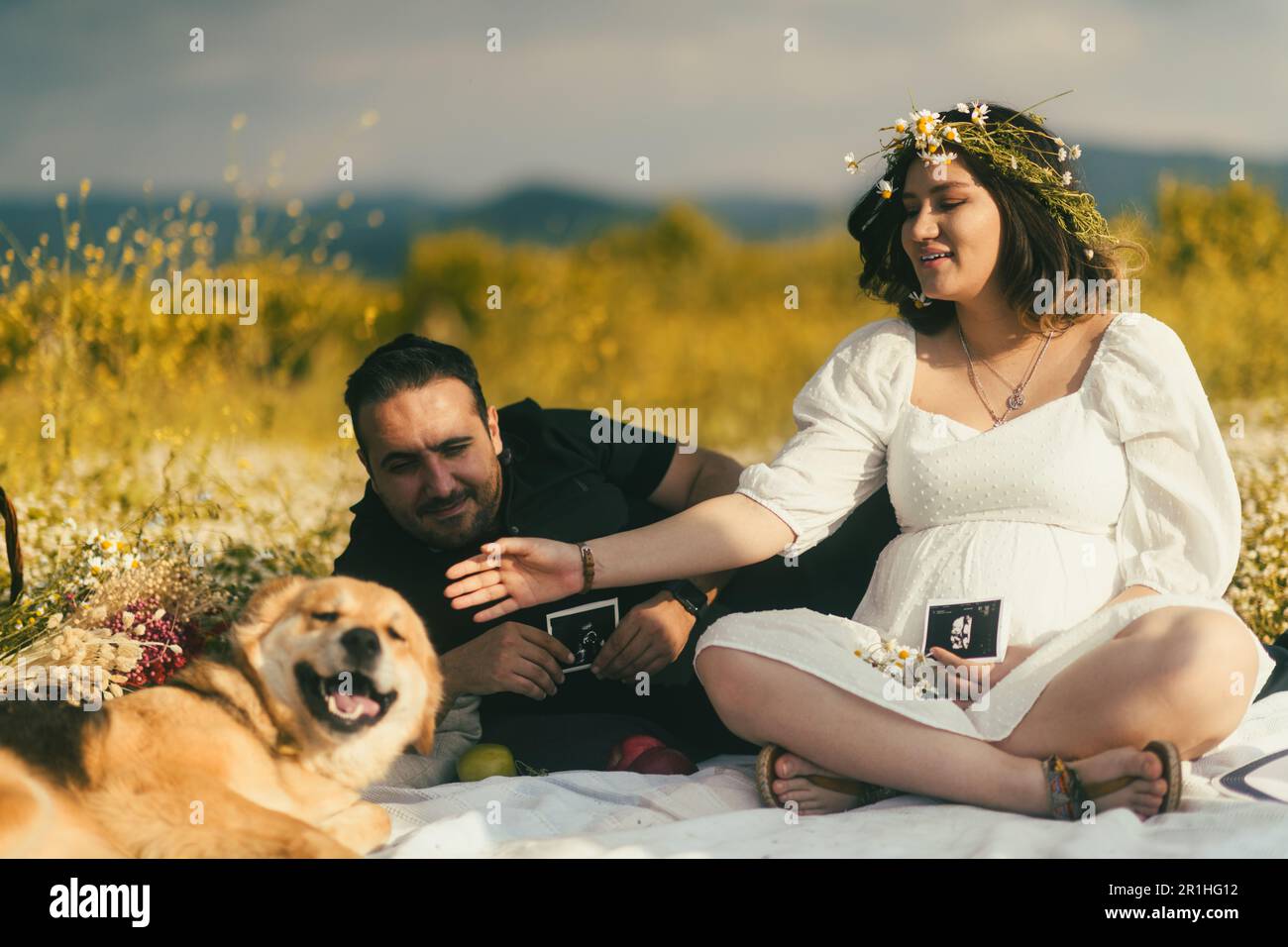 Sur une couverture de pique-nique blanche, une femme enceinte et son mari partagent un moment de tendreté avec l'image échographique de leur bébé, tandis que leur sympathique Golden Retr Banque D'Images