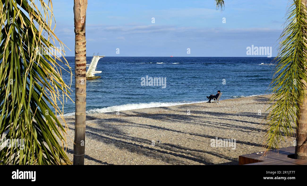 Côte de mer avec plage de galets. Côte d'hiver déserte. Une tour de plongée est visible dans la mer. Un homme assis sur une chaise de plage est assis sur la rive Banque D'Images