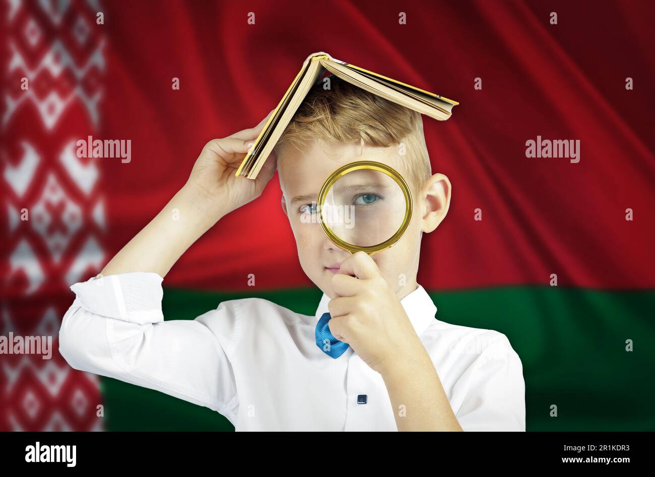 Un garçon avec un livre sur sa tête regarde à travers une loupe sur le fond du drapeau biélorusse. Concept d'éducation. Banque D'Images