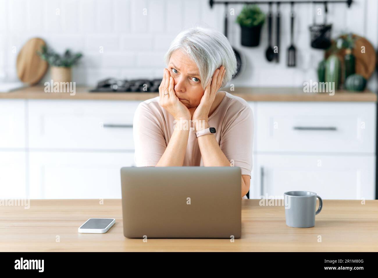 Perplexe, exténuée, exténuée, femme à cheveux gris caucasien, retraité, assis à une table avec un ordinateur portable dans la cuisine, se sent surtravaillé et stressé, fatigue de travailler sur un ordinateur portable, regarde malheureusement l'appareil photo Banque D'Images