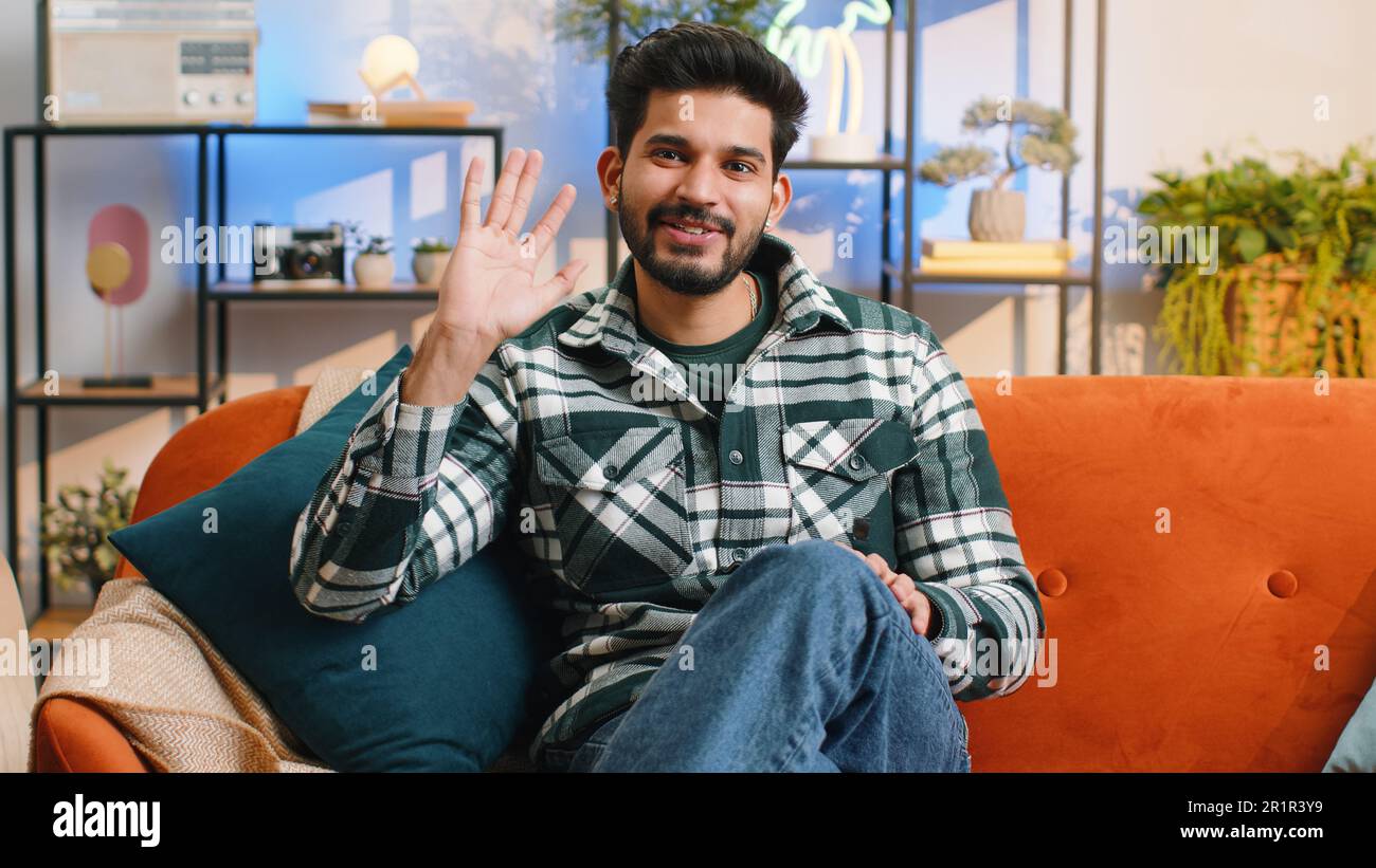 Bonjour. Homme indien souriant amical à l'appareil photo et en agitant les mains en signe de bonjour, salut, salutation ou au revoir, accueillant avec une expression accueillante à la maison. Un homme hindou assis sur un canapé dans la salle de séjour Banque D'Images