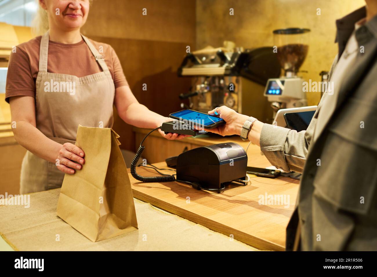 Prise de vue d'un employé de boulangerie mûr tenant un sac en papier avec des aliments à emporter ou des pâtisseries et un terminal de paiement pendant que le client paie pour l'achat Banque D'Images