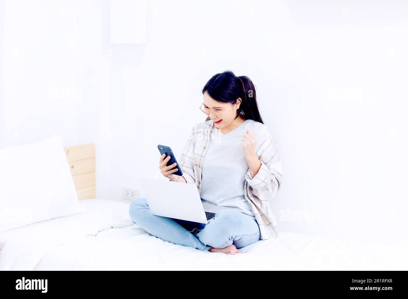 Femme souriante assise sur son lit à pieds croisés à l'aide d'un téléphone portable et d'un ordinateur portable Banque D'Images