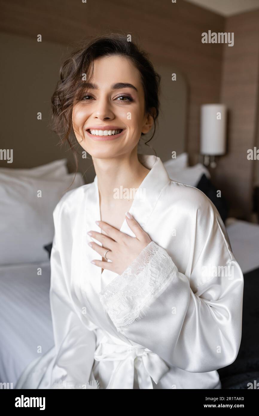 jeune mariée heureuse avec cheveux brunette et anneau d'engagement sur le  doigt souriant tout en étant assis dans un peignoir en soie blanche et  regardant l'appareil photo dans la chambre d'hôtel le