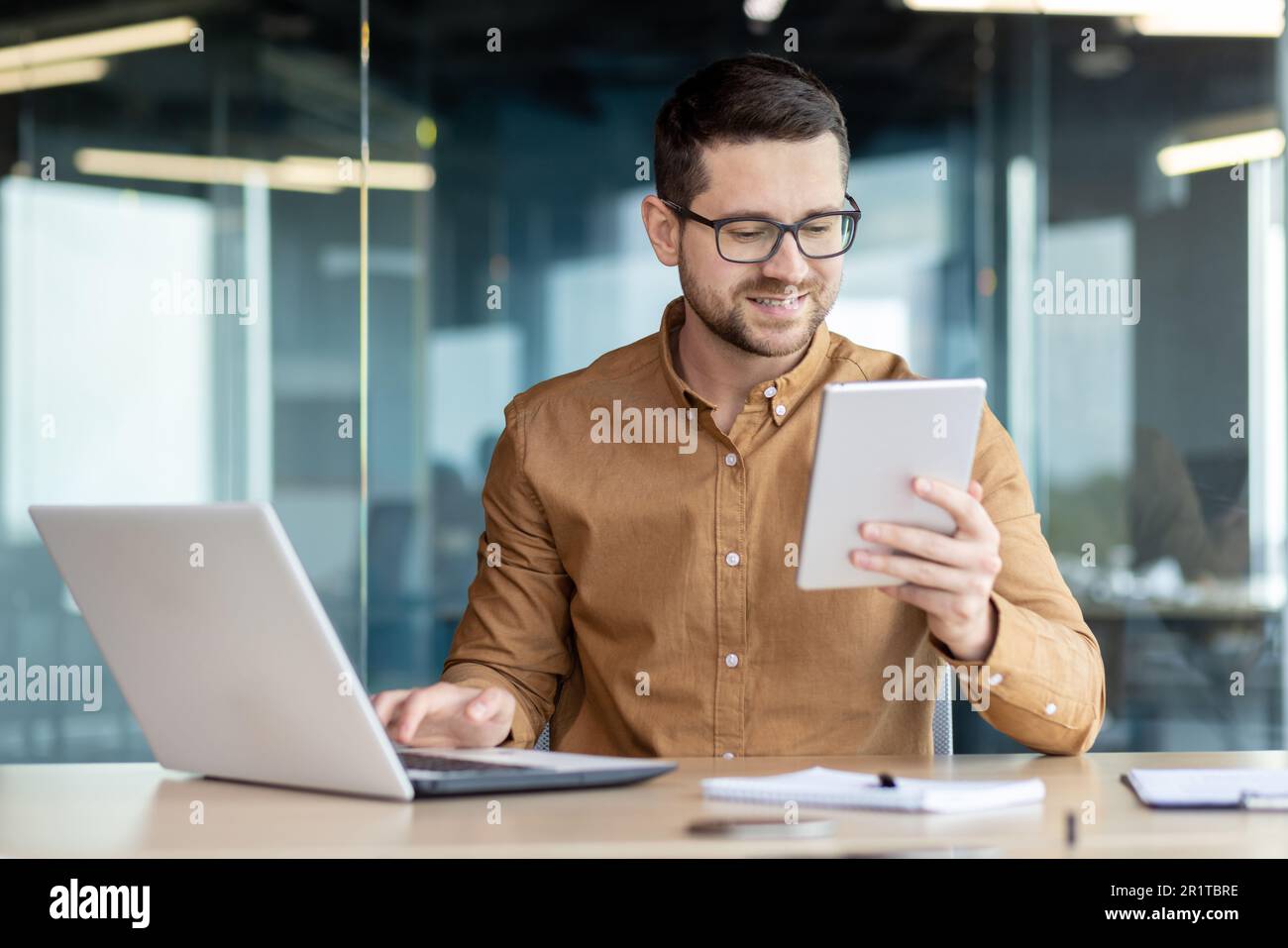 Un jeune homme travaille dans un centre de bureau, s'assoit à une table avec un ordinateur portable, tient une tablette dans la main et parle lors d'un appel vidéo avec le sourire. Banque D'Images