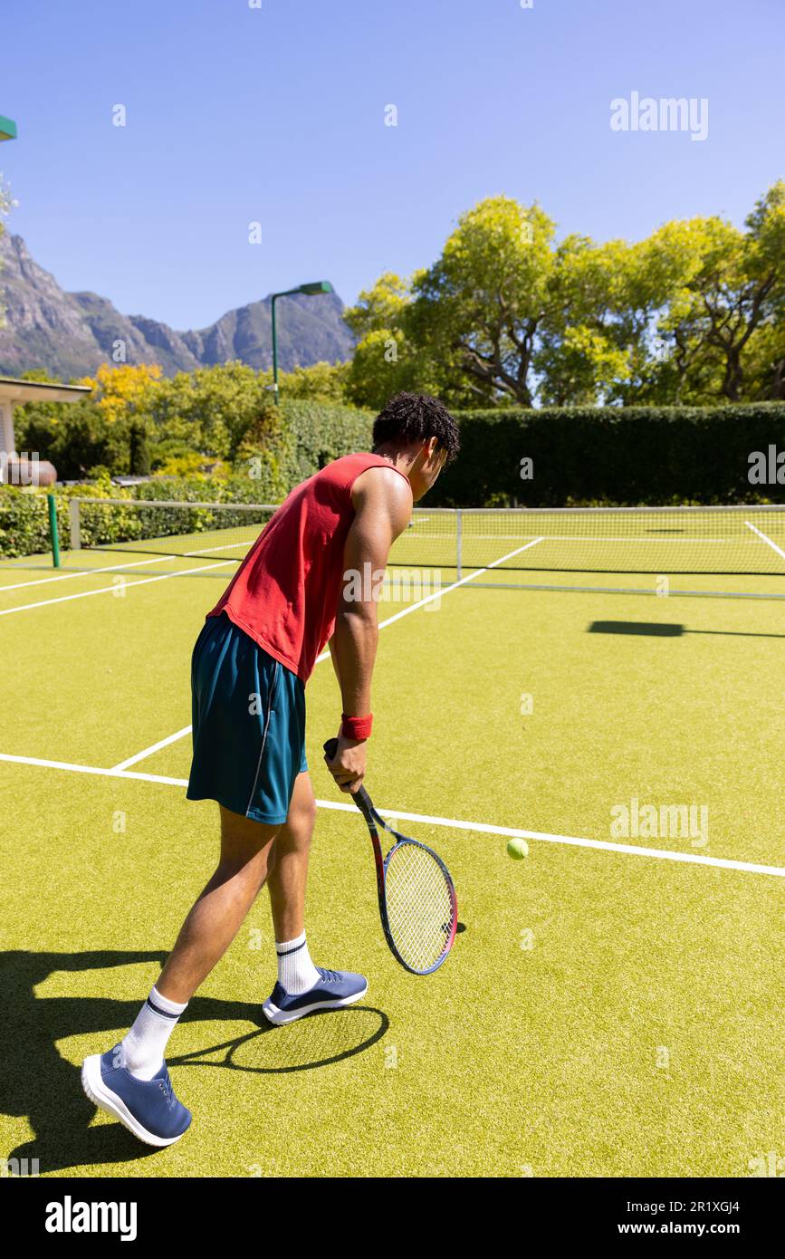 Homme biracial avec une raquette de tennis rebondissant pour servir sur un court de tennis extérieur ensoleillé, espace copie Banque D'Images