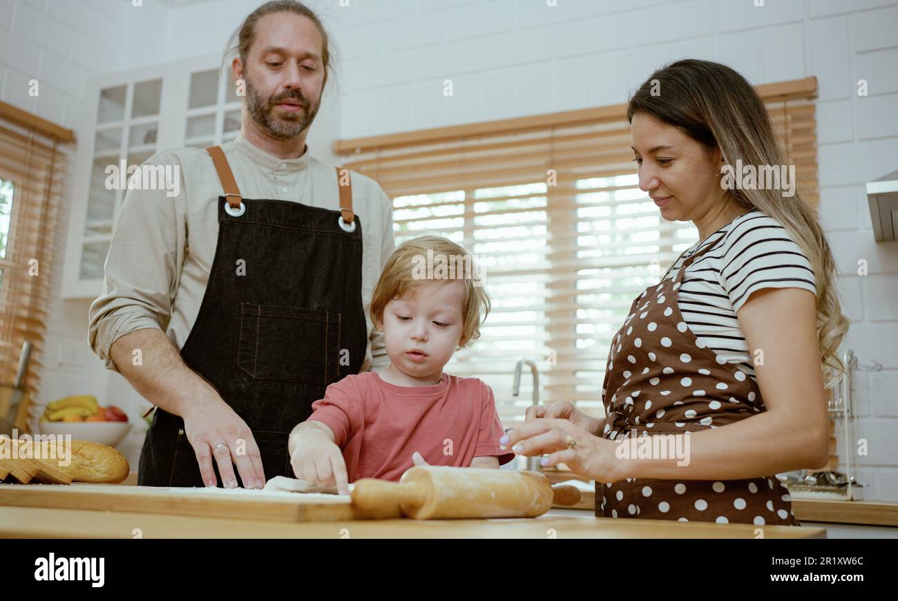 Le père et la mère enseignent à bébé le pétrissage de la pâte sur le comptoir de cuisine à la maison. Les parents et le garçon gamin apprécient et s'amusent à cuisiner à l'intérieur ensemble. Banque D'Images