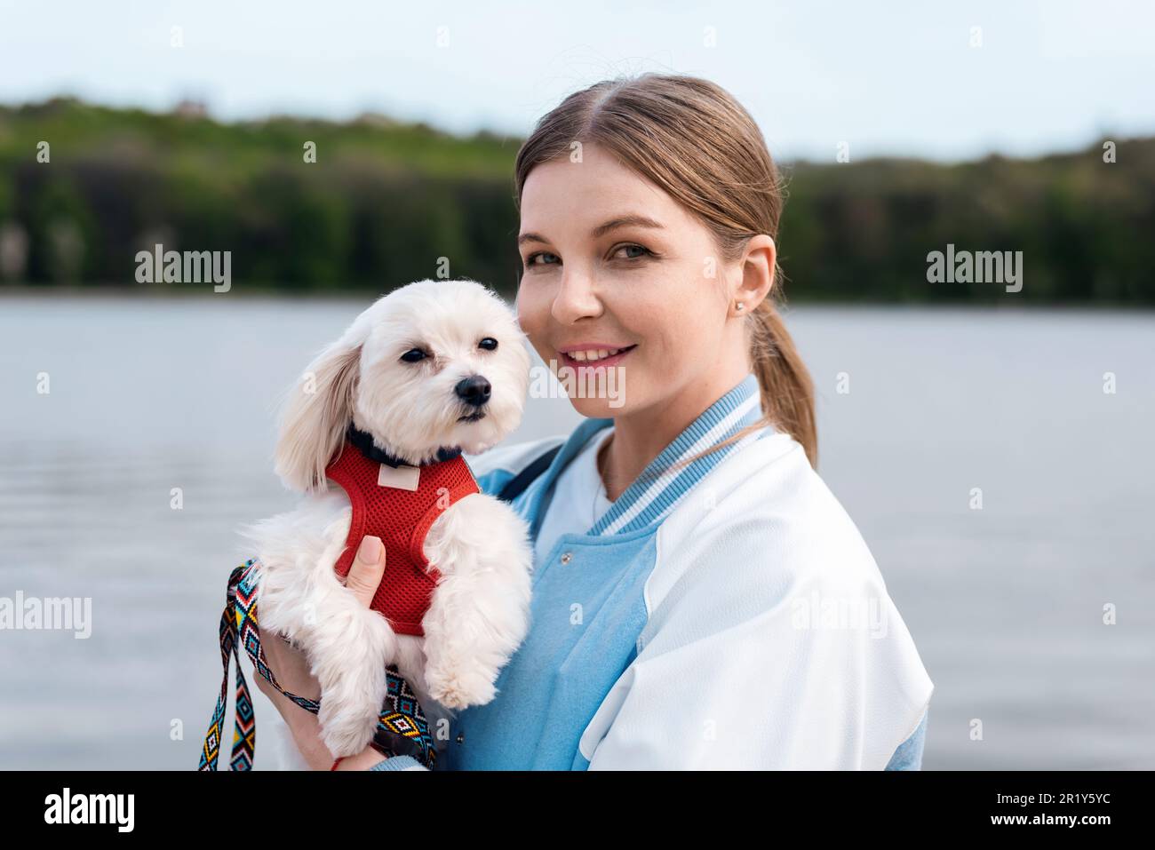 Vue d'une femme blonde tenant son chien maltais à la fourrure blanche et regardant dans l'appareil photo, lac sur le fond Banque D'Images