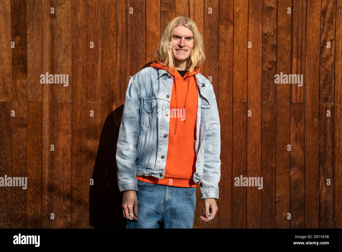Joyeux positif millenny hipster gars avec de longs cheveux blonds debout à l'extérieur souriant à la caméra Banque D'Images