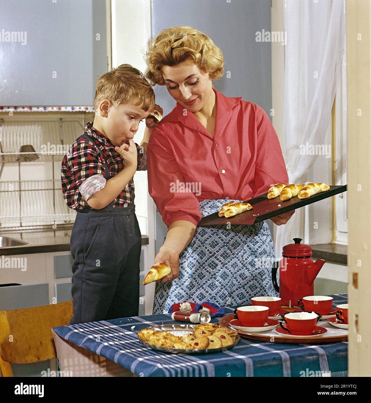 Cuisson dans le 1950s. Une mère avec son fils dans la cuisine. Elle a des petits pains et des gâteaux fraîchement cuits au four et le garçon semble comme s'il aimait l'odeur agréable d'eux. Une image typique de 1950s avec les vêtements de la mère, les tasses et la boîte, l'intérieur de la cuisine et les couleurs. Suède 1958 Conard Banque D'Images